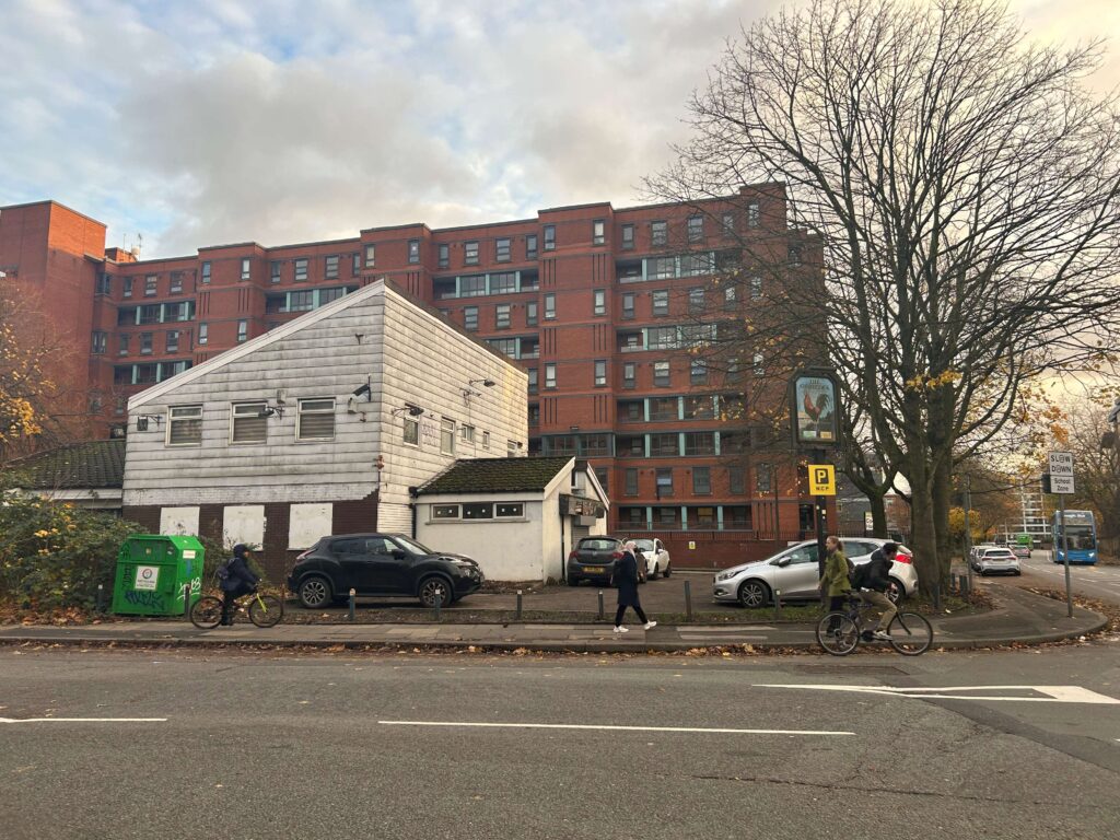 A photo of the abandoned Gamecock Pub, a dirty white building with a  large red brick building behind it. The windows are shuttered, and a large sign with a cockerel stands outside.