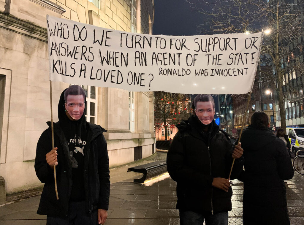 Ronaldo Johnson's family holding a white homemade banner, which reads: "Who do we turn to for support or answers when an agent of the state kills a loved one? Ronaldo was innocent."
The banner is being held by two boys wearing masks of Ronaldo's face. They are standing at a candelit vigil outside Friends Meeting House, and it is dark outside. 