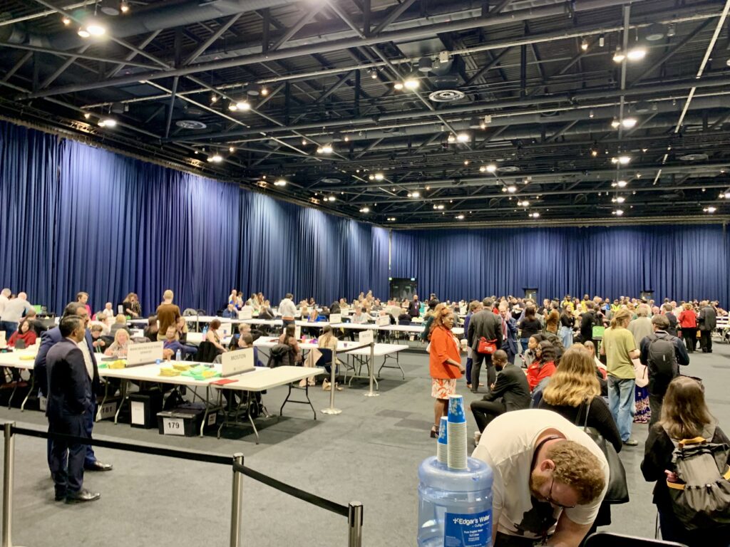 A large room full of people inside the Manchester Conference Hall during the election count. It is well lit and crowded.