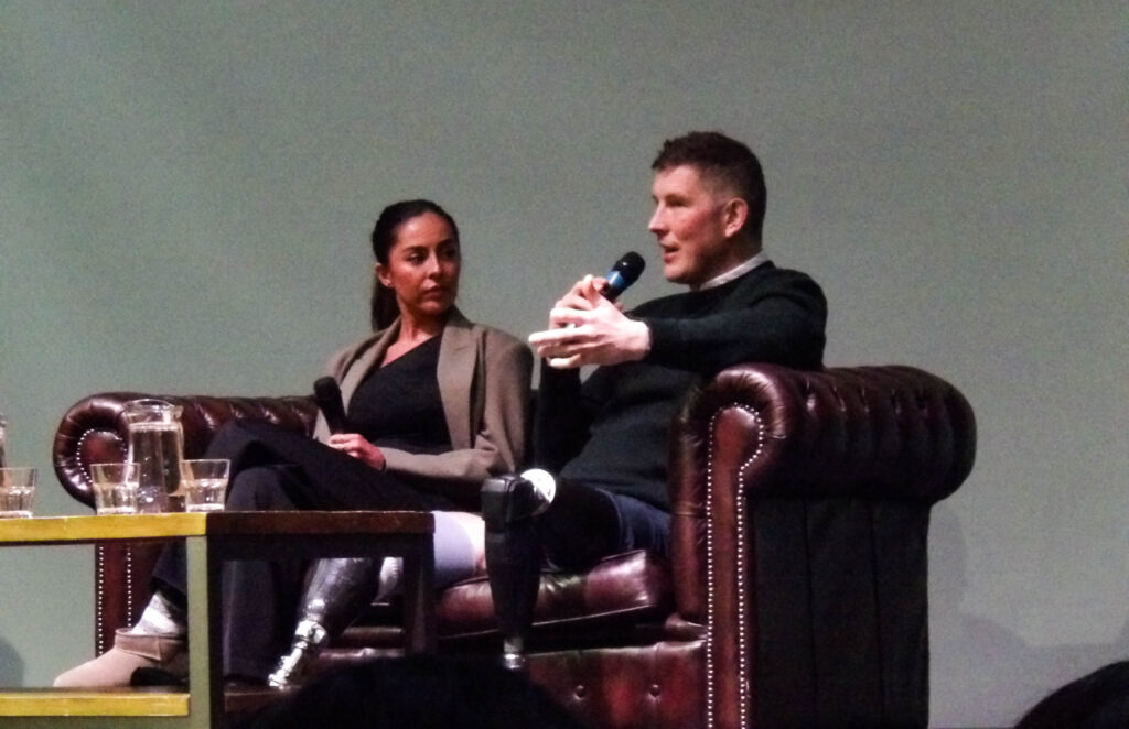 A brown woman and white man sit next to each other on a leather sofa, both holding microphones. The woman (left) is wearing a black shit and grey blazer, while the man wears a green jumper. The man has two black prosthetic legs.
