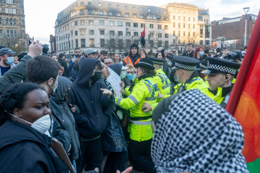 A group of police officers in high-vis jackets stand opposite a crown of protesters wearing face masks.
