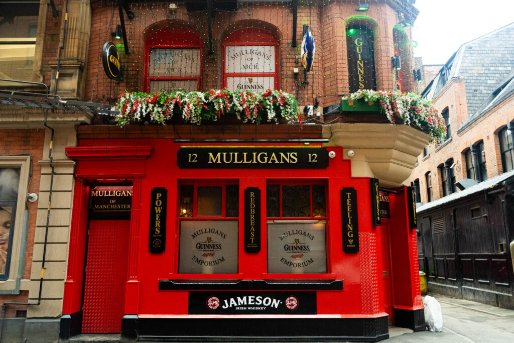 The side of Mulligans Irish Bar. It is a brick building, and the lower half is built entirely of bright red painted metal. A black sign that reads 'MULLIGANS' is above two darkened windows, and below a long basket of fake red and white flowers. The doors are shuttered. The top half of the building is covered in switched-off fairy lights.