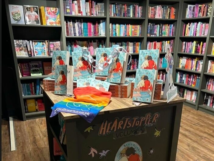 A table inside Queer Lit. The table is filled with hardback copies of Alice Oseman's Heartstopper graphic novel. There is a rainbow tote bag with the Queer Lit logo on display alongside them. In the background are rows of bookshelves.