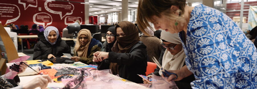 Dr Lynn Setterington, a white woman with short brown hair and a blue checked button-up shirt, leans over a table on the right side of the image. She is sharing the table with a crowd of women, and the table is covered in scraps of cloth, scissors, and other tools. They are hard at work stitching something together in a large, open-plan studio.