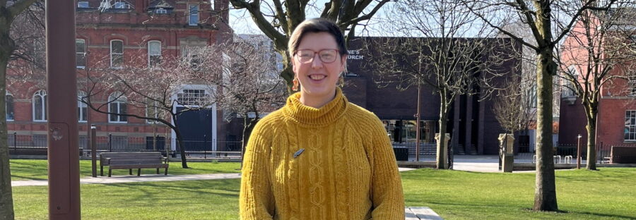 Iris Skipworth, a white woman with short blonde hair, glasses, yellow jumper, and blue jeans, stands with her legs crossed and hands clasped, smiling a the camera. She is leaning against a picnic table in All Saints Park, with trees and grass in the background. It is a sunny day.