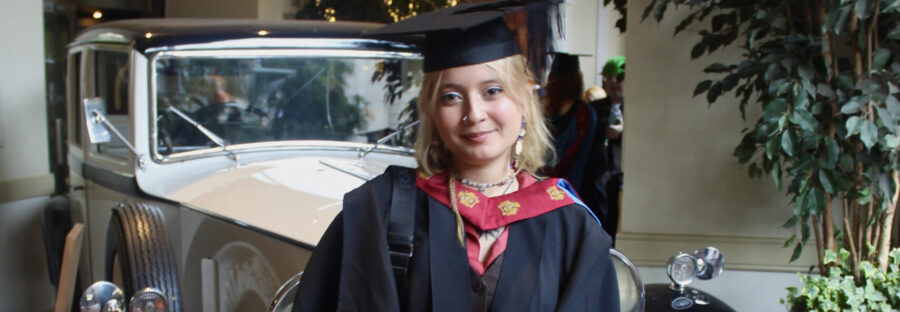 Alisson Ponce is a young white woman with wavy, bleach-blonde hair. She is wearing a red and white graduate gown and cap, and standing in front of a vintage car. She is holding a diploma and smiling.