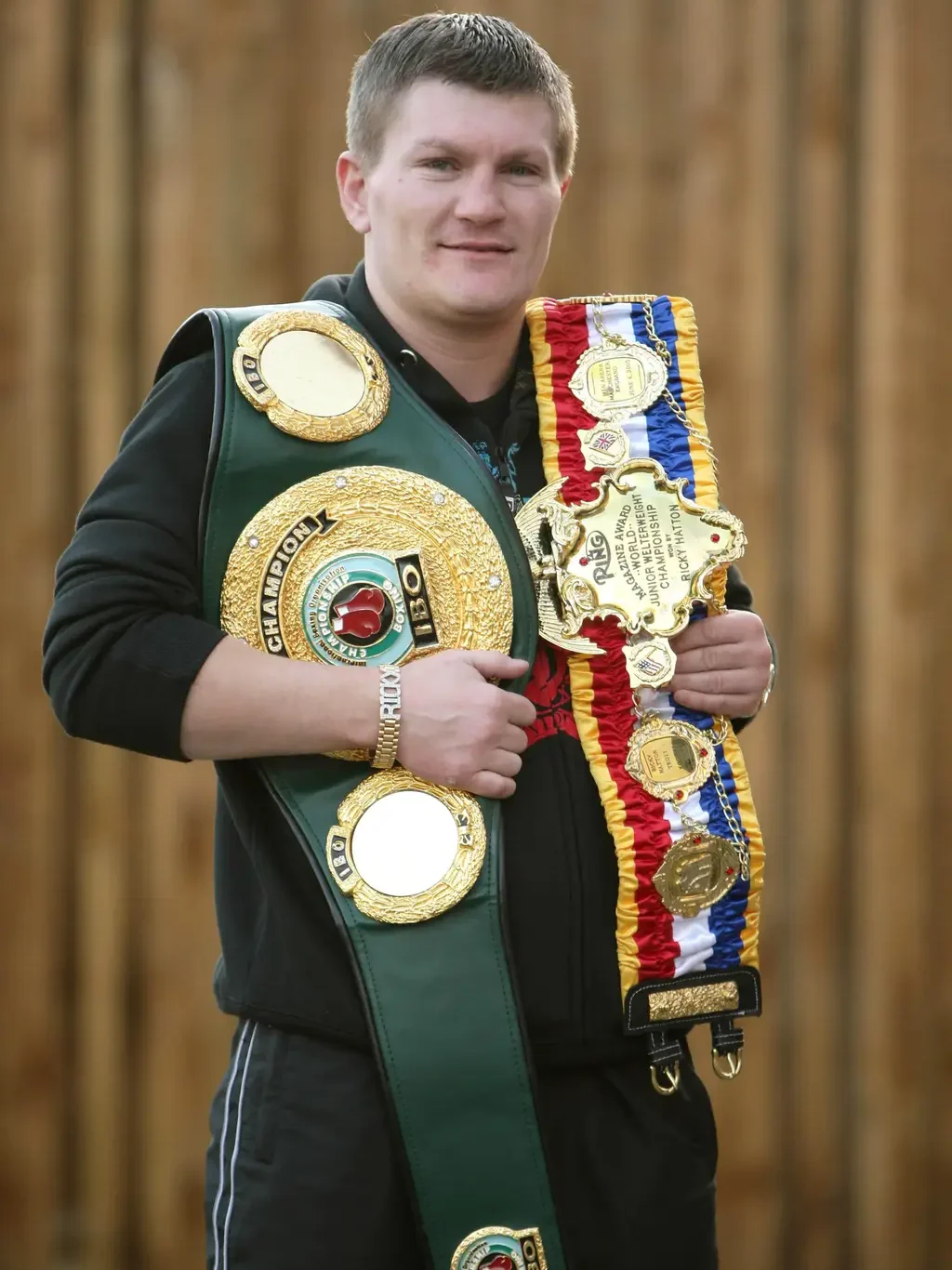 Ricky Hatton is a white man with short blond hair wearing a black shirt and trousers. He holds two champion belts over his shoulders, both with large golden pieces. He is smiling. 