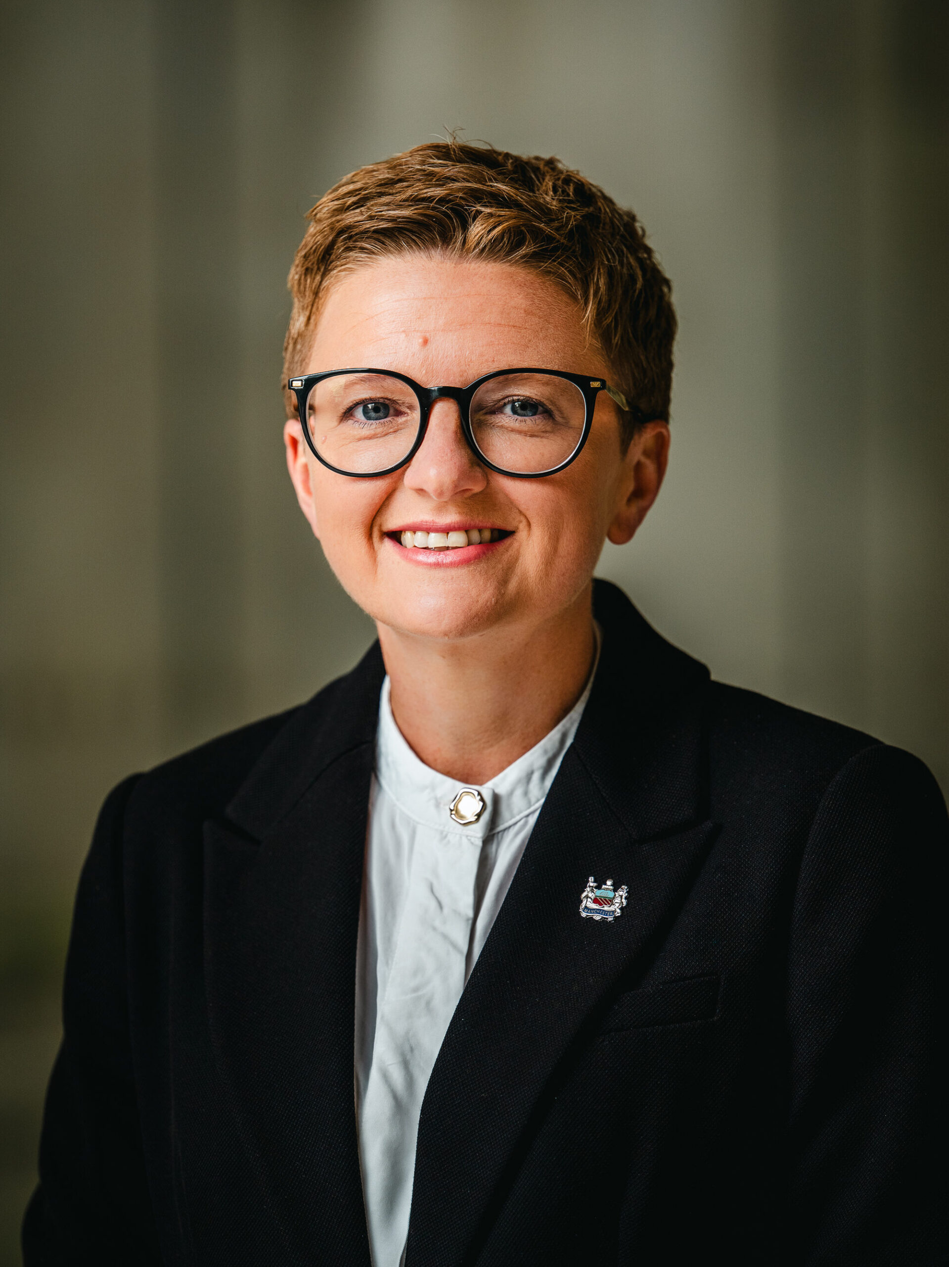 Bev Craig is a white woman with very short blonde hair. She is wearing large glasses and a black suit with a Manchester City Council pin badge on the lapel. She is smiling at the camera.