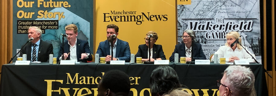 Three white men and three white women sit at a long black table, each with a microphone and mug. The tablecloth has the Manchester Evening News logo in yellow. Behind the people are three banners for the Manchester Evening News. From left to right is Marlon West, Richard Filpatrick, Phil Eckersley, Bev Craig, Geraldine Coggins, and Sian Astley. All are in formal wear. A crowd is visible in the foreground, as the image was taken from behind them.