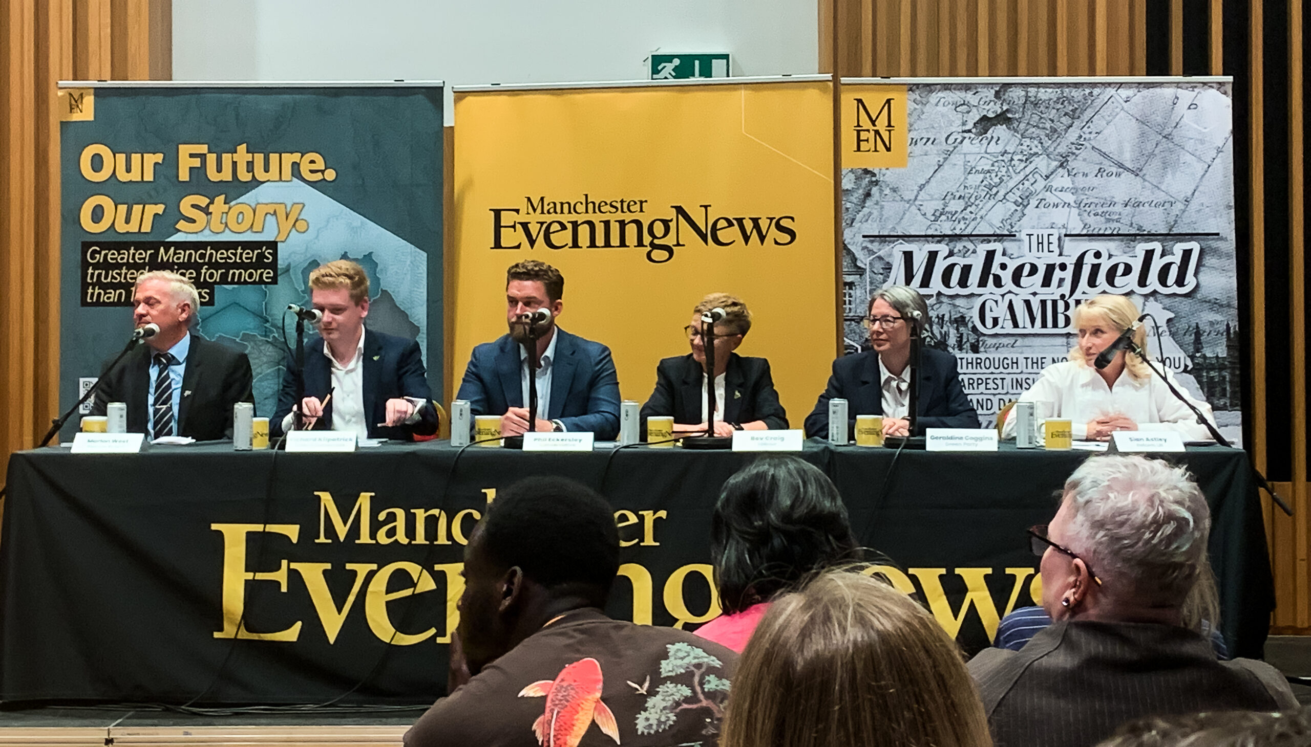 Three white men and three white women sit at a long black table, each with a microphone and mug. The tablecloth has the Manchester Evening News logo in yellow. Behind the people are three banners for the Manchester Evening News. From left to right is Marlon West, Richard Filpatrick, Phil Eckersley, Bev Craig, Geraldine Coggins, and Sian Astley. All are in formal wear. A crowd is visible in the foreground, as the image was taken from behind them.