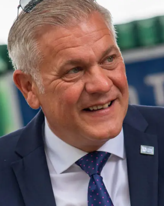 Marlon West is an older white man with slicked back grey hair. He is wearing a navy suit and tie, with a Restore Britain badge on the lapel. He is talking and looking away from the camera.