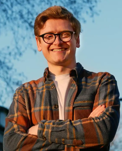 Richard Kilpatrick is a young white man with ginger hair and round glasses. He is wearing a white T-shirt and orange flannel jacket, and has his arms folded. He is smiling into the camera