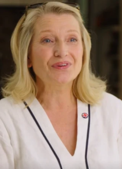 Sian Astley is an older white woman with long blonde hair and lipstick. She wears a white dress shirt and a Union Jack pin. She is mid speech and looking away from the camera.