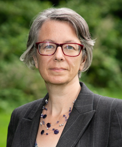 Geraldine Coggins is a white woman with ear-length grey hair. She is wearing a red pair of glasses, a black sit jacket, and a necklace of purple marble pebbles. She is half smiling into the camera.